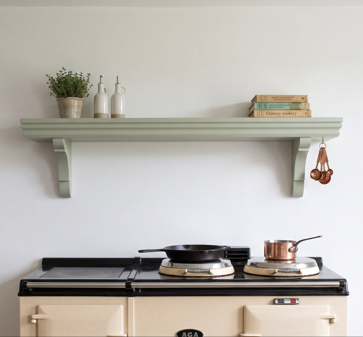 A cream-colored Aga stove with a black cooktop sits beneath a pale sage green decorative shelf mounted on a white wall. The shelf holds a small potted plant, two white bottles, and a stack of cookbooks, with copper measuring cups hanging on the right side.