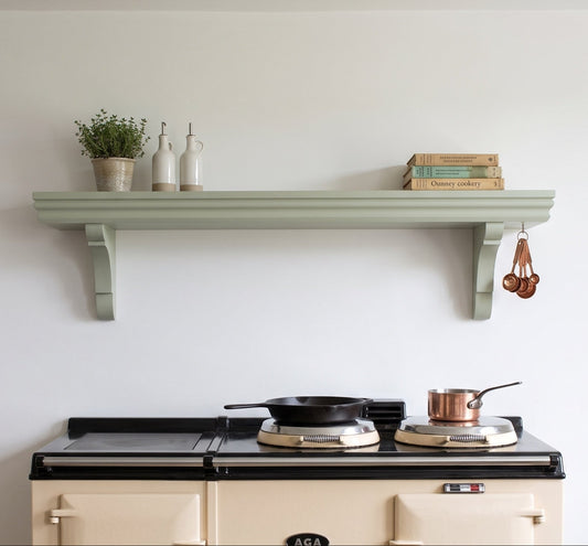 A cream-colored Aga stove with a black cooktop sits beneath a pale sage green decorative shelf mounted on a white wall. The shelf holds a small potted plant, two white bottles, and a stack of cookbooks, with copper measuring cups hanging on the right side.